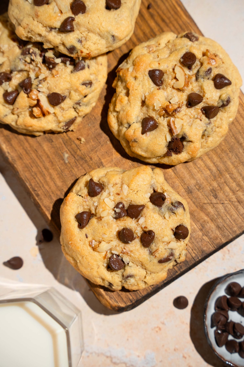 A wooden board with Neiman Marcus chocolate chip cookies. The board is on a tan counter with a small plate of chocolate chips and a glass of milk.