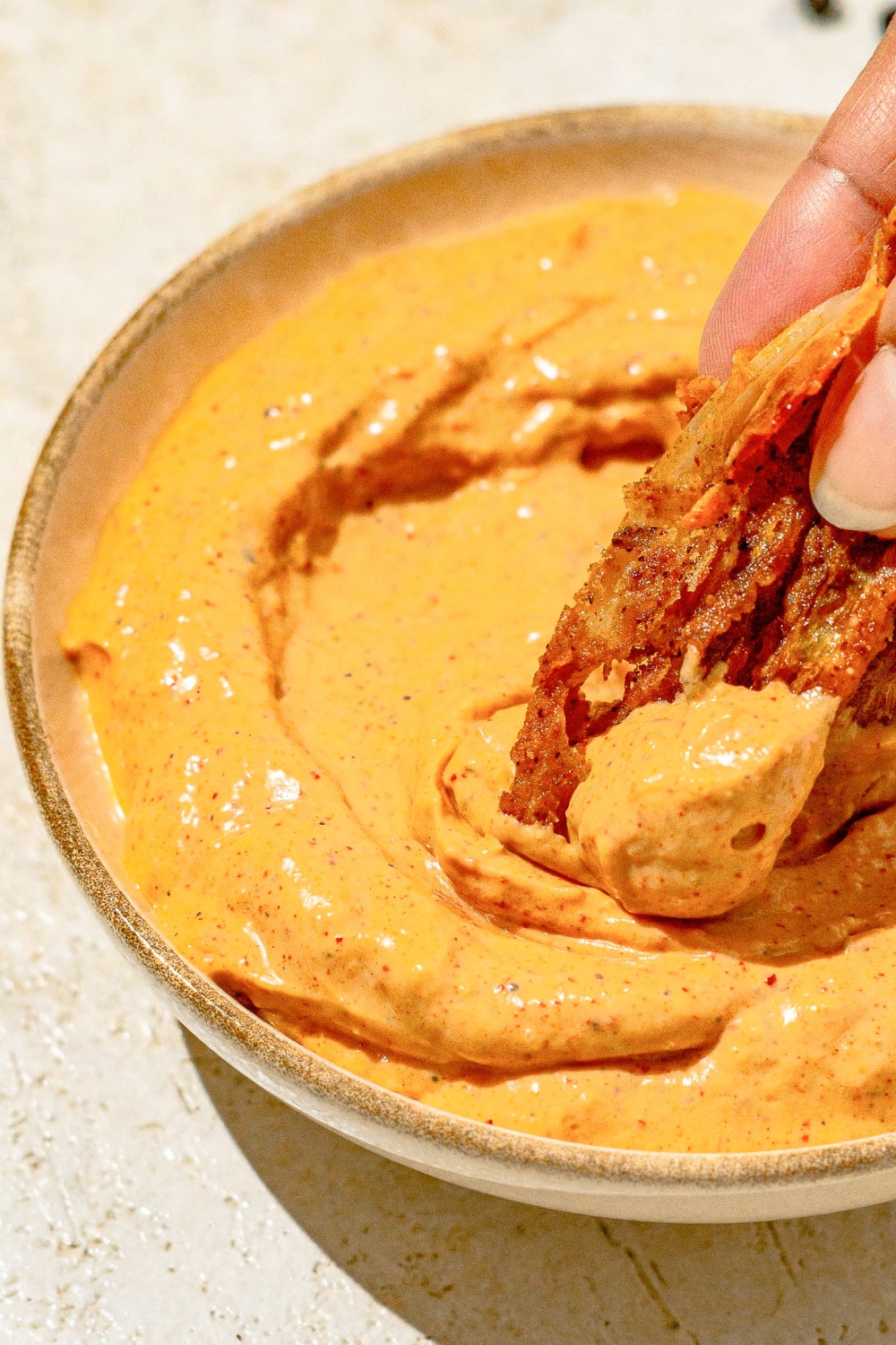 A hand dipping a piece of Outback bloomin onion into a bowl of onion sauce. The bowl is on a tan counter.