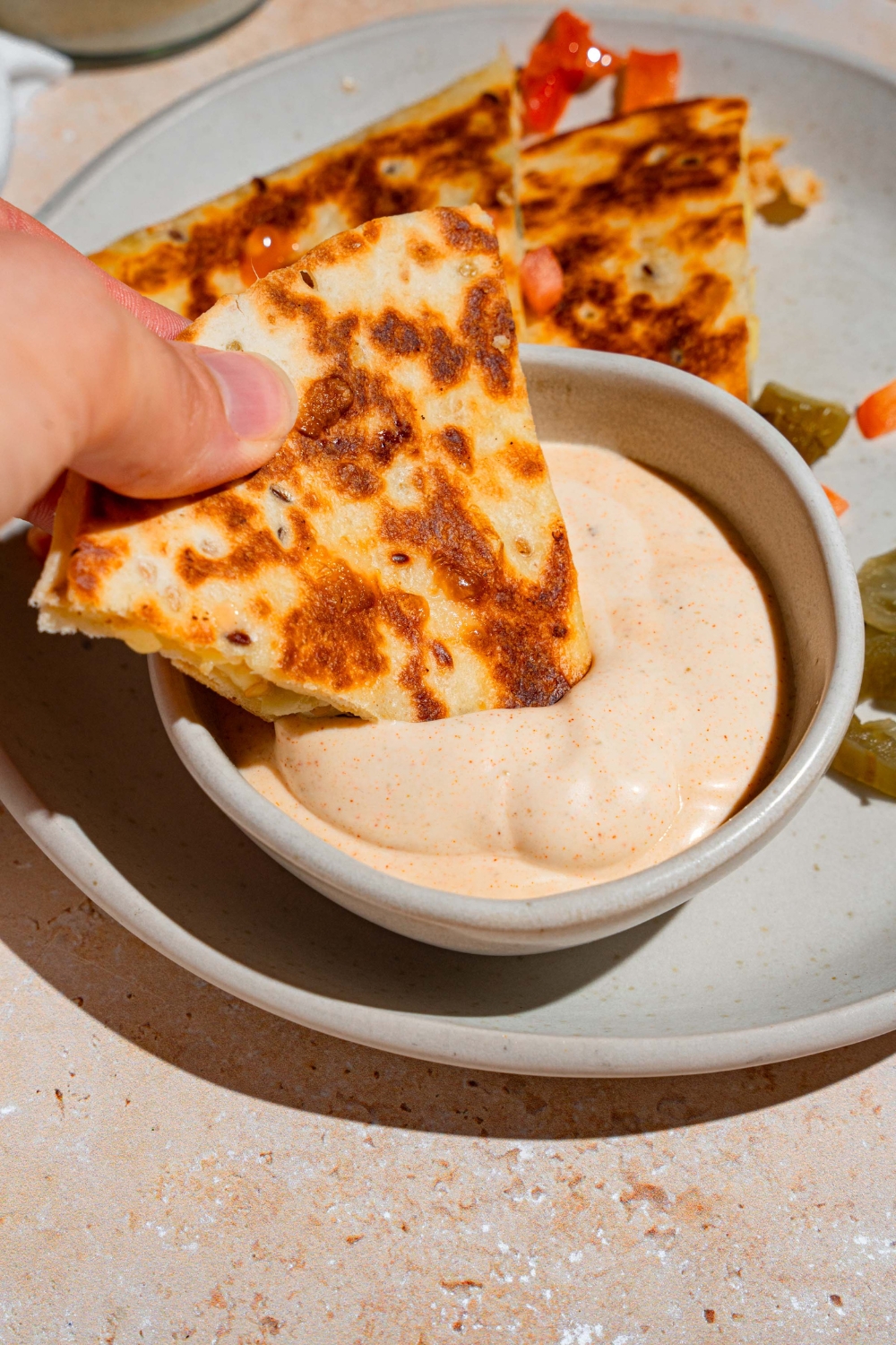 A hand dipping a slice of quesadilla into a bowl of Taco Bell quesadilla sauce. The bowl is on a white plate with a quesadilla garnished with peppers and pickled jalapenos.