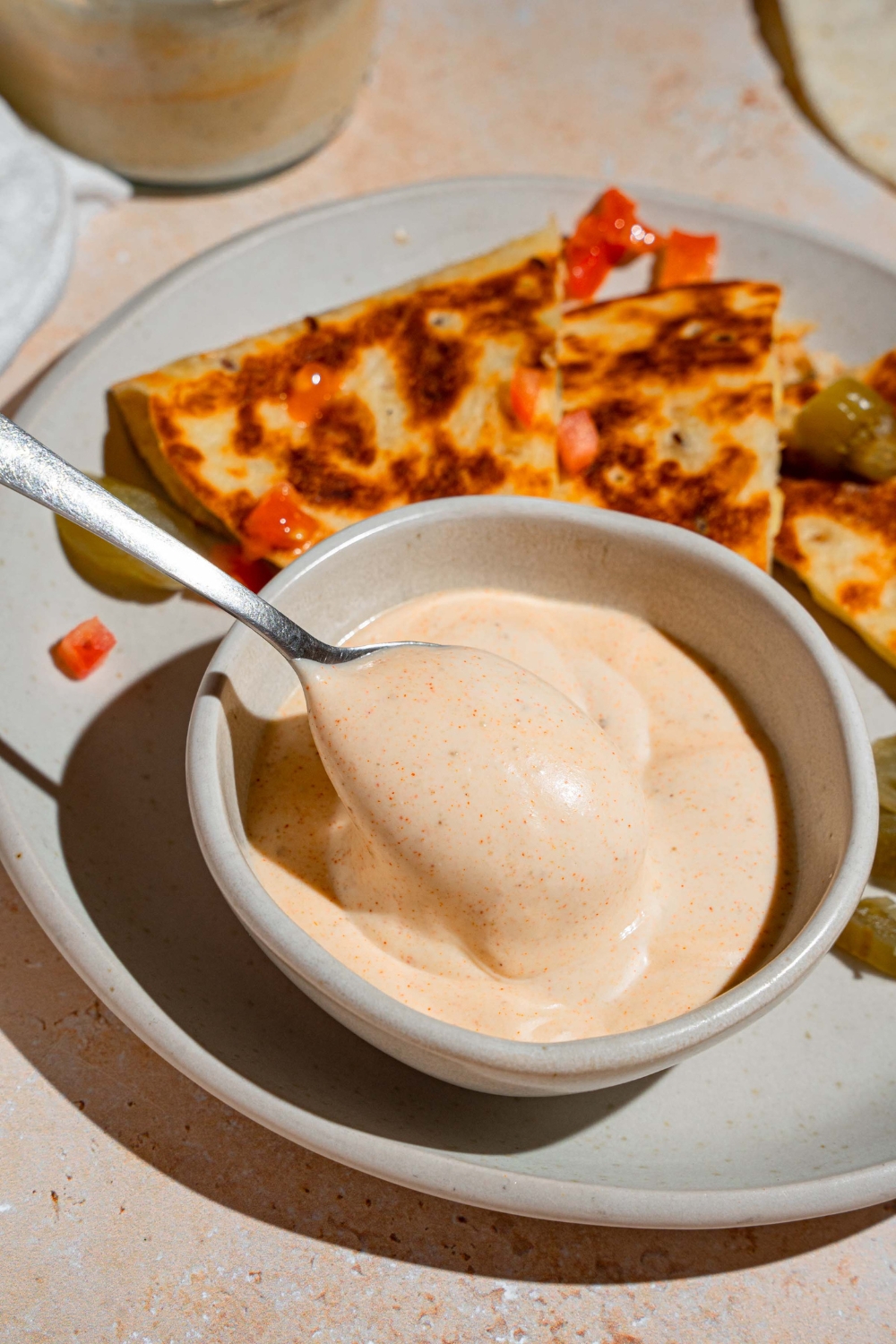 A spoon dipping into a bowl of Taco Bell quesadilla sauce. The bowl is on a white plate with a quesadilla garnished with peppers and pickled jalapenos. The plate is on a tan counter.