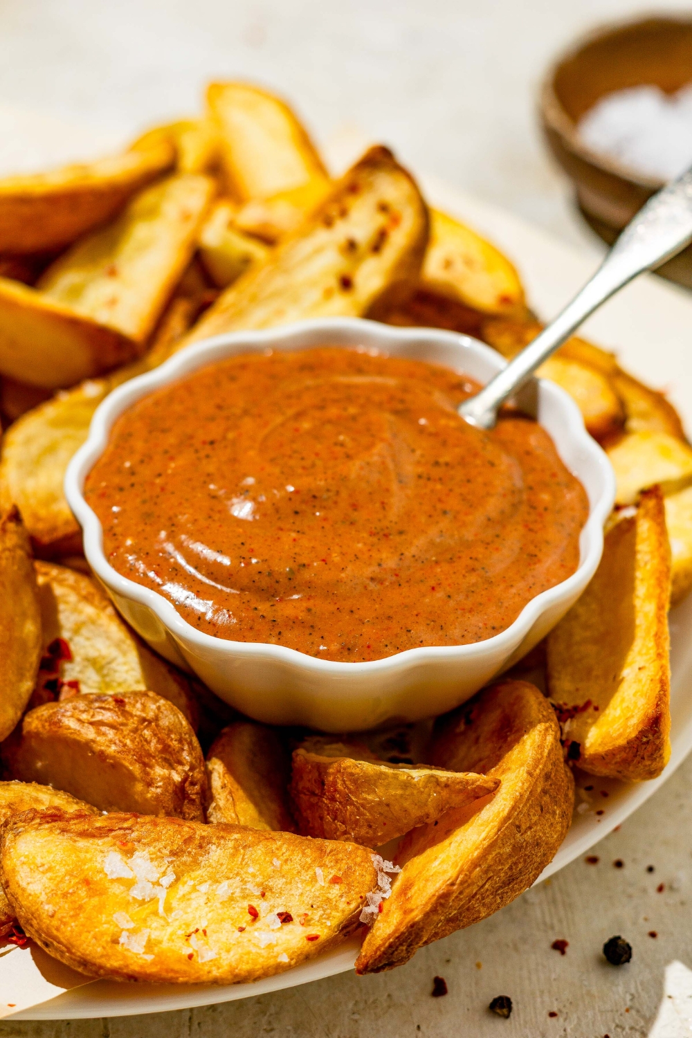A small bowl of copycat Zaxby's sauce on a plate with wedge fries. There is a spoon in the sauce. The plate is on a tan counter with a small bowl of salt.
