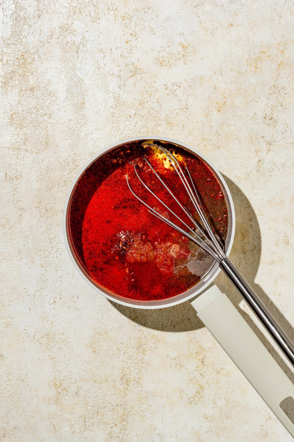 A whisk mixing ingredients to make Carolina BBQ sauce in a small pot. The cup is on a tan counter.