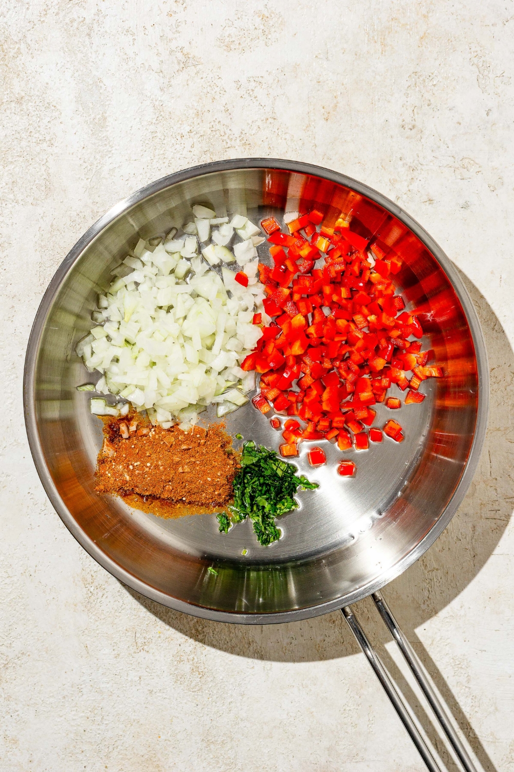 A skillet with diced onion, diced peppers, seasonings, and cilantro. The skillet is on a tan counter.