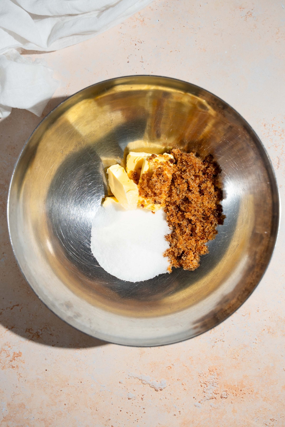 A mixing bowl with butter, sugar, and brown sugar. The bowl is on a tan counter with a white cloth napkin.