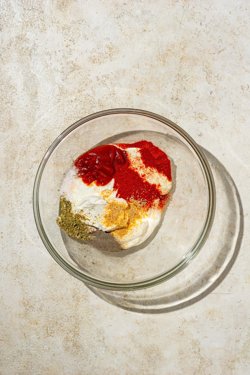 A glass bowl with ingredients to make copycat Outback bloomin onion sauce including ketchup, mayo, sour cream, and seasonings. The bowl is on a tan counter.