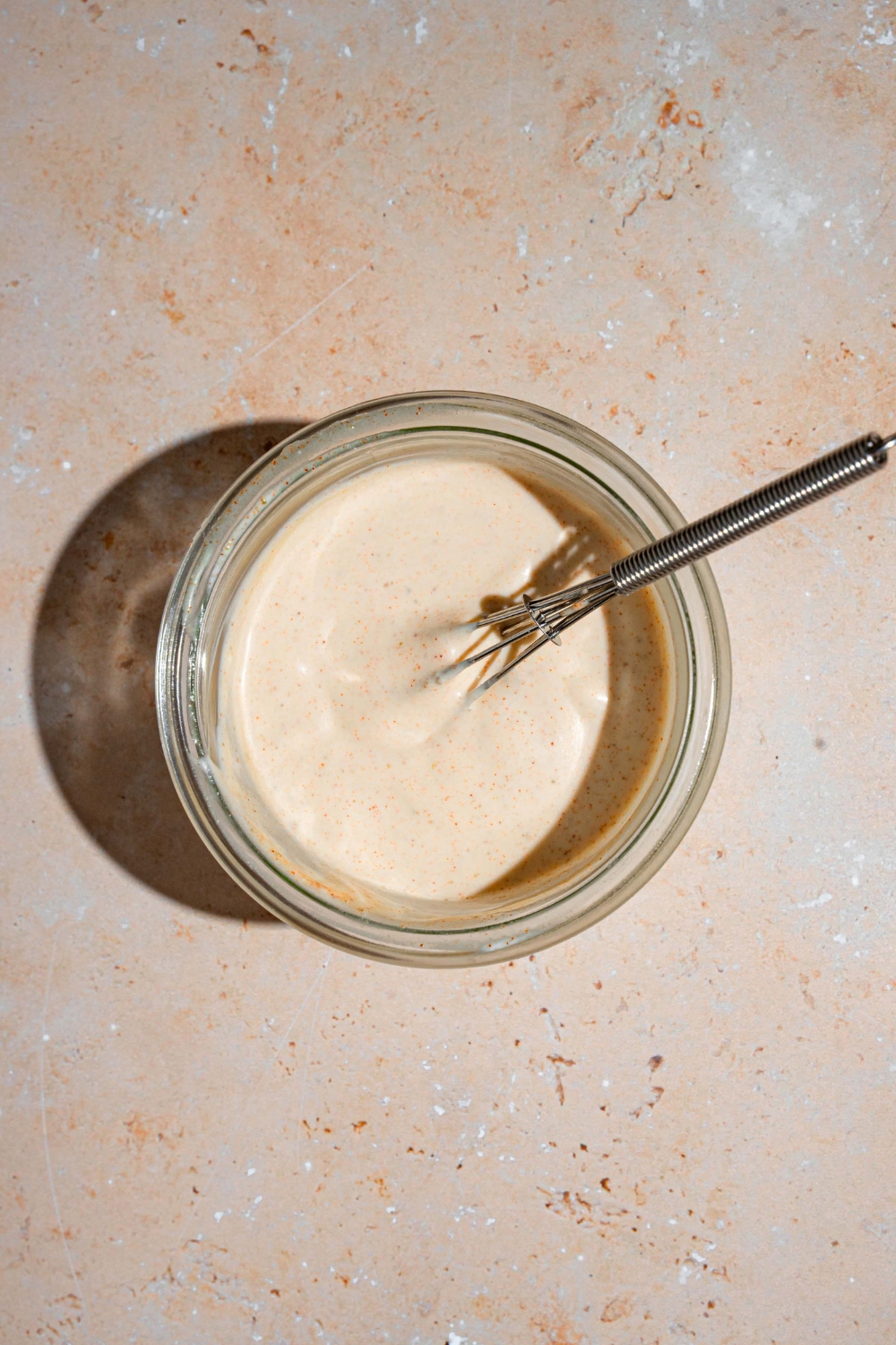 A glass jar filled with Taco Bell quesadilla sauce. There is a small whisk in the jar. The jar is on a tan counter.
