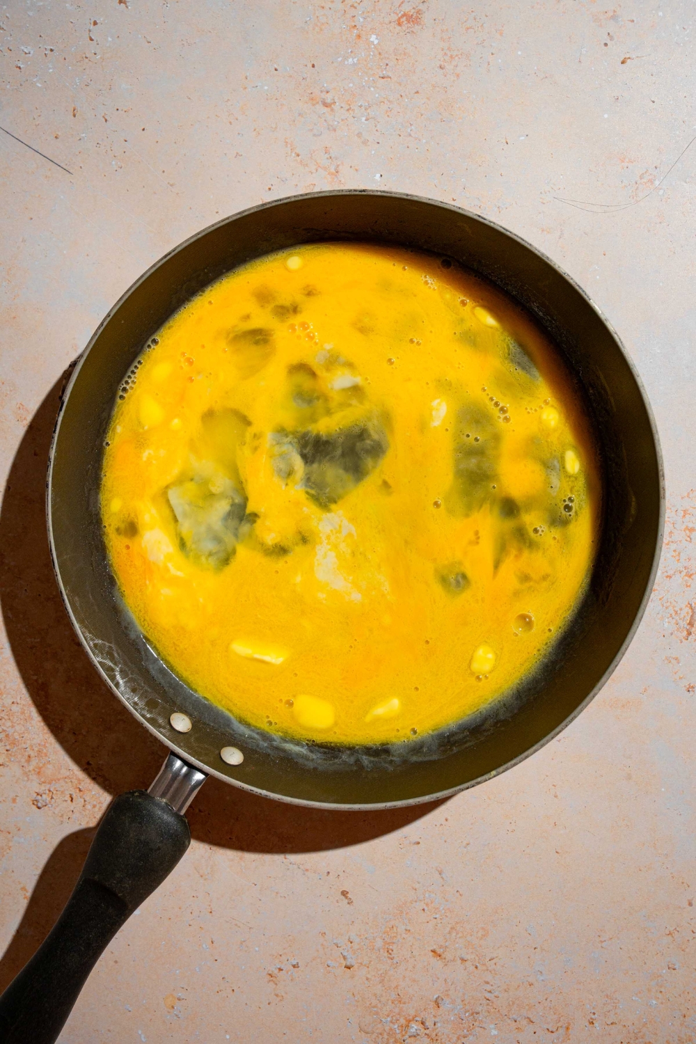 A skillet with scrambled uncooked eggs with butter. The skillet is on a tan counter.