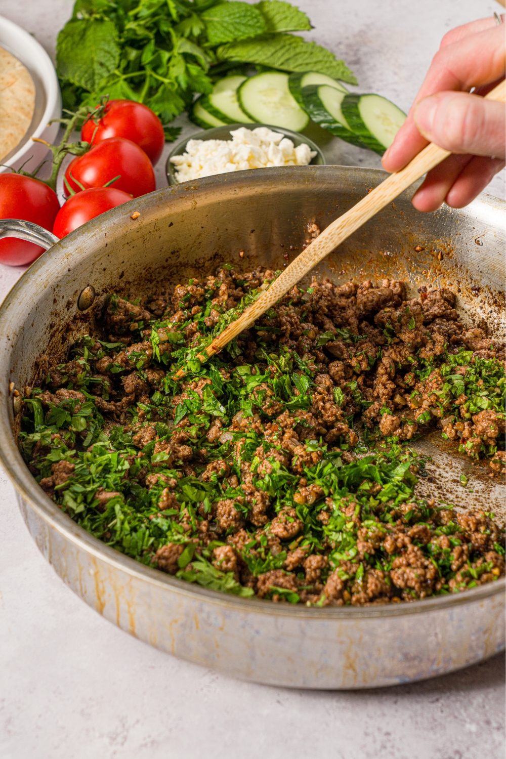 A skillet with a spoon mixing seasoned ground lamb with fresh parsley. The skillet is on a white counter with fresh tomatoes, cucumber, and a bowl of feta.