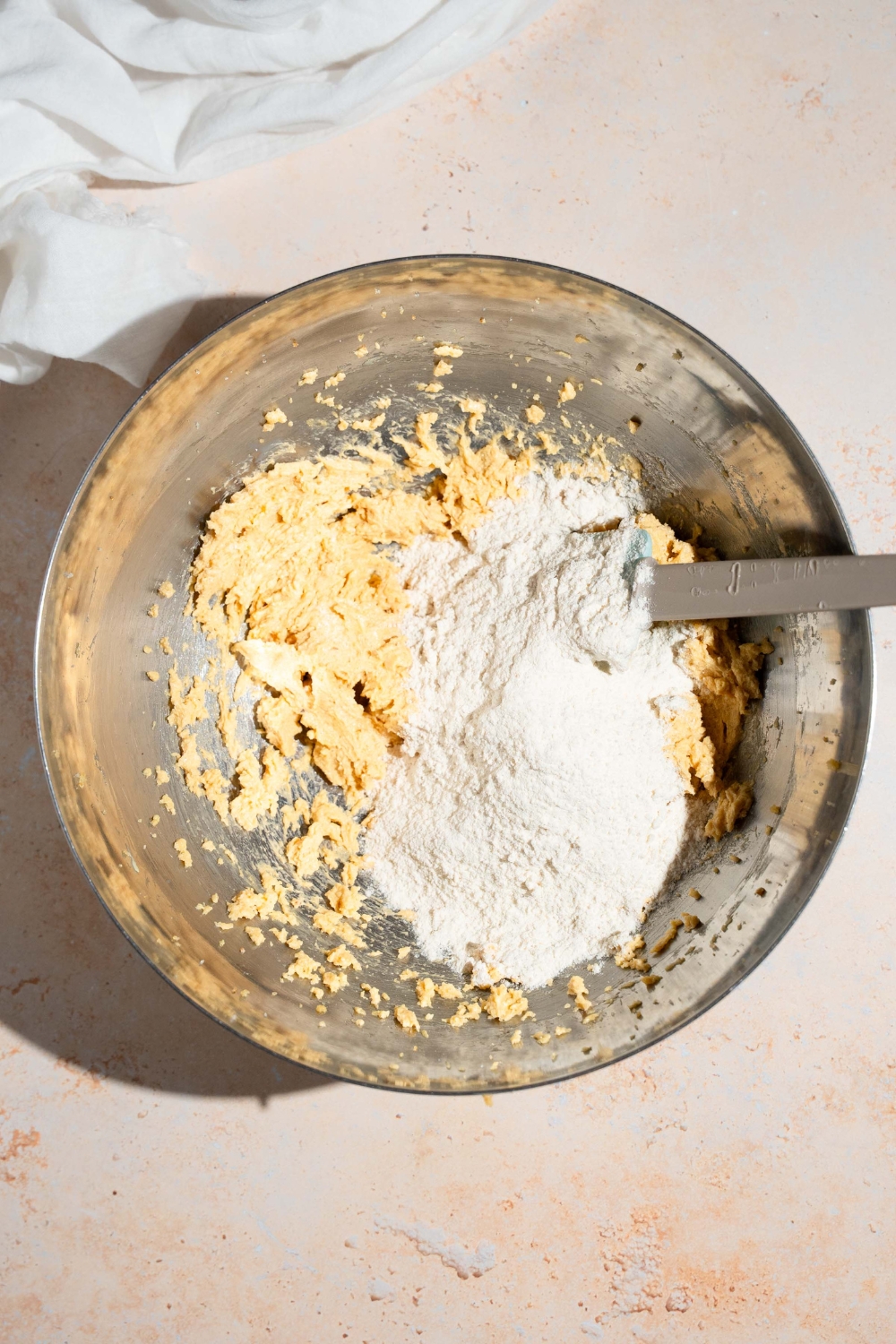 A mixing bowl with a spatula combining a butter mixture with dry ingredients. The bowl is on a tan counter with a white cloth napkin.