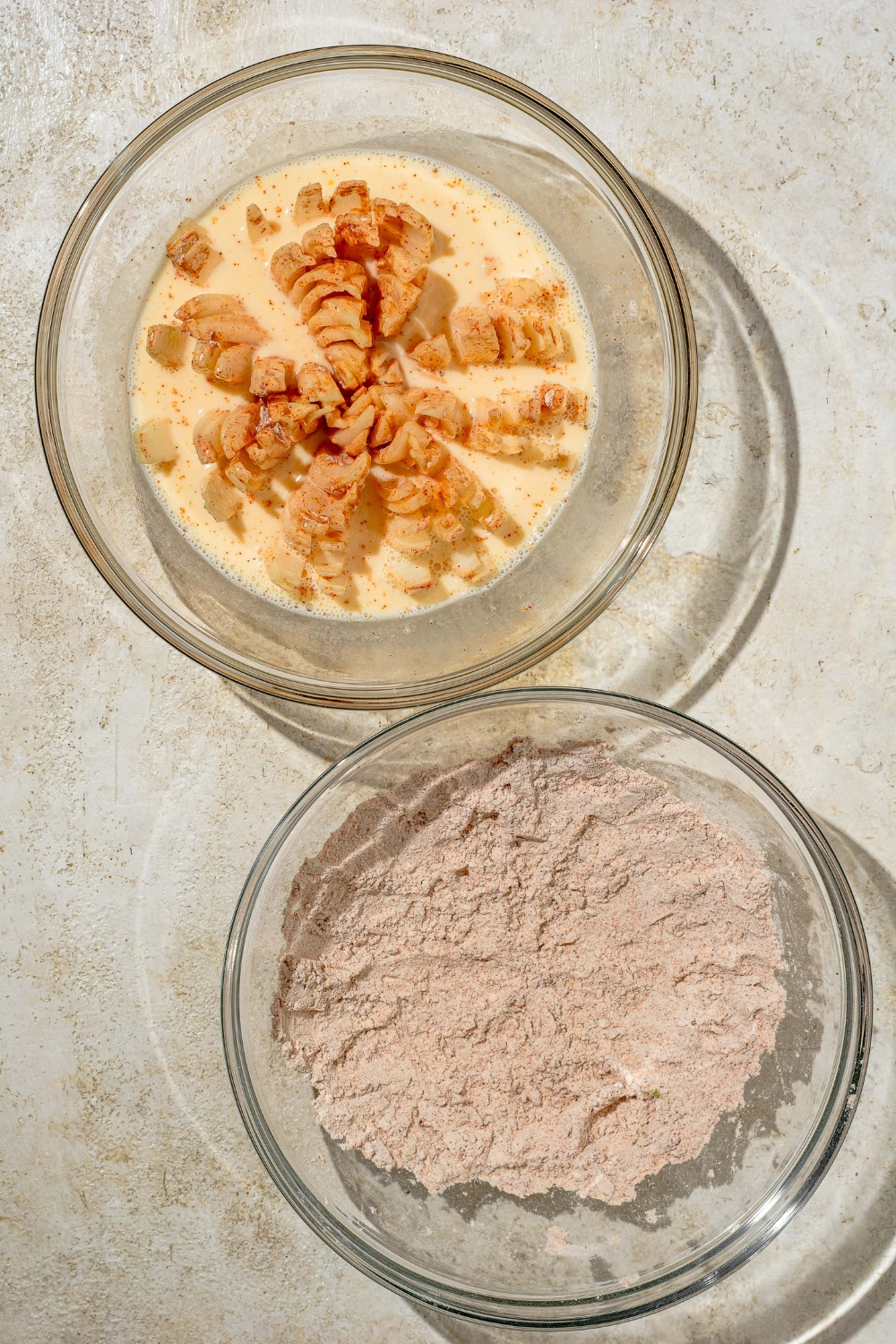 A glass bowl with a flour and seasoning mixture next to an additional glass bowl with a fried bloomin onion sitting in an egg mixture.