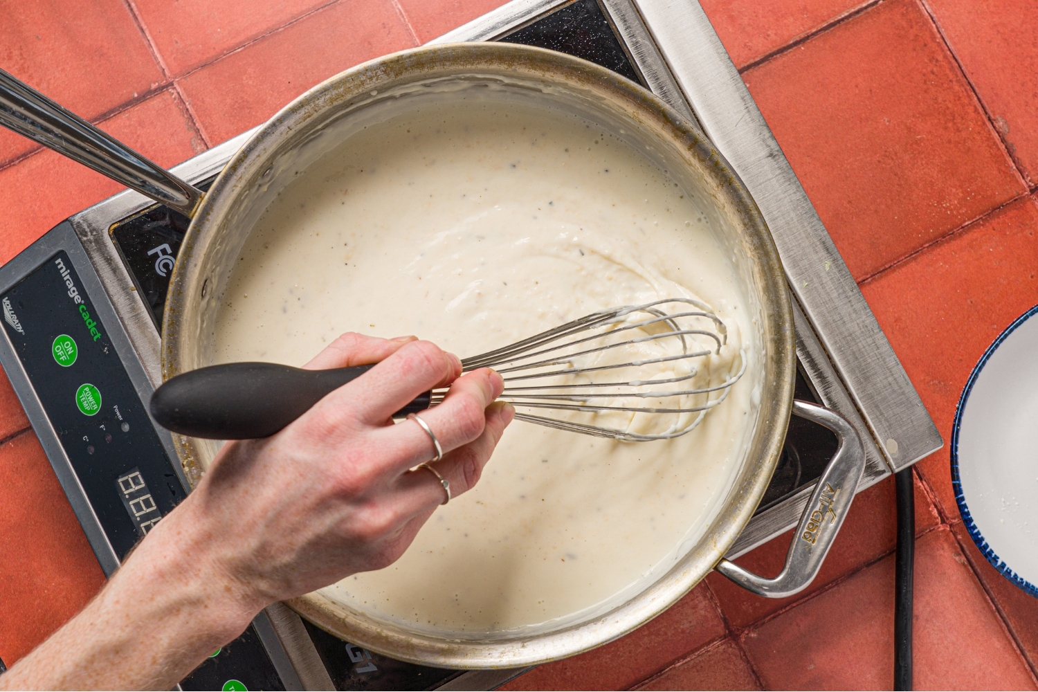 A skillet with alfredo sauce cooking over a burner. There is a hand stirring the sauce with a whisk. The burner is on a brick counter.