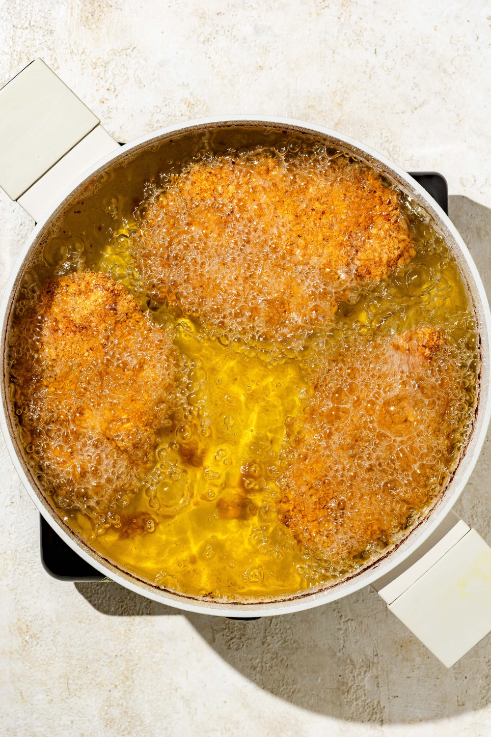 A deep pot with several pieces of breaded chicken frying in oil. The pot is on a tan counter.