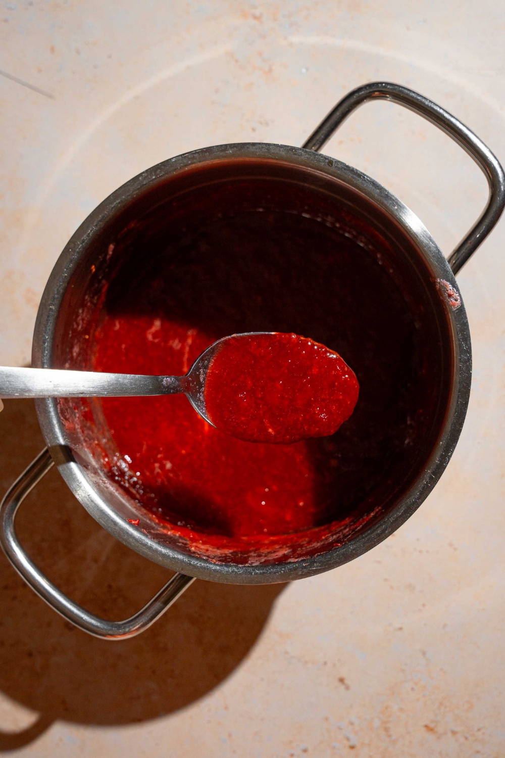 A stock pot with cooked Christmas jam. A spoon is dipping into the jam. The pot is on a tan counter.