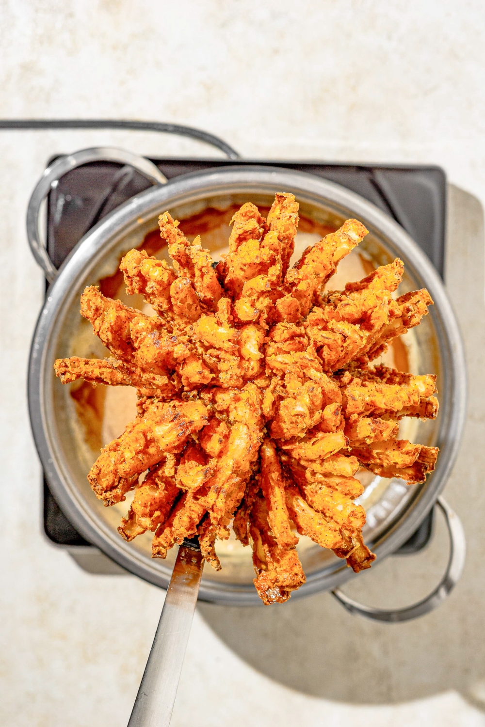 A mesh spoon holding a fried Outback bloomin onion over a pot on a burner. The burner is on a tan counter.