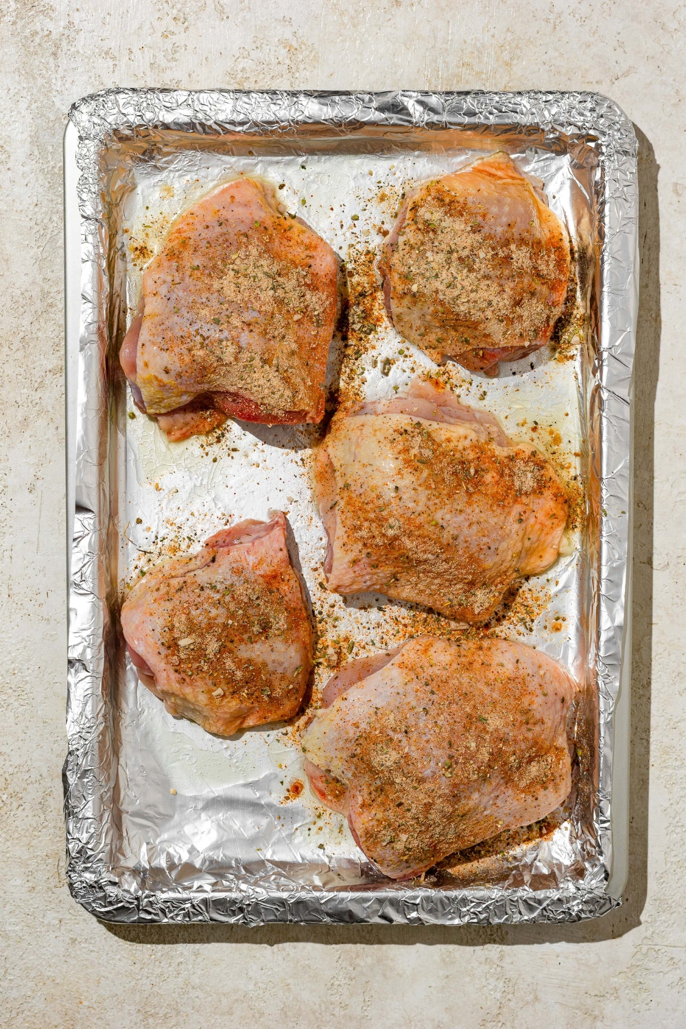 A baking sheet lined with aluminum foil with chicken thighs marinating. The sheet is on a tan counter.
