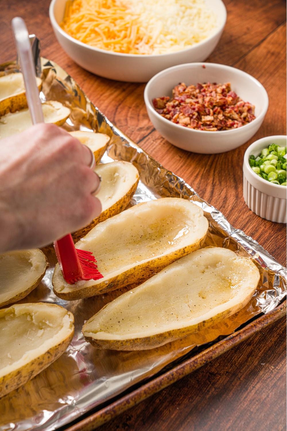 A baking sheet lined with foil with prepared potato skins. A hand is brushing butter on the insides of the potato skins. The tray is on a wood counter with small bowls of toppings.