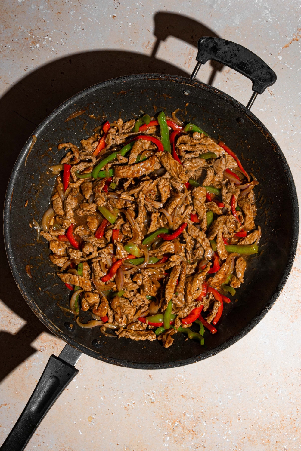 A wok with cooked pepper steak tossed with sliced peppers. The wok is on a tan counter.
