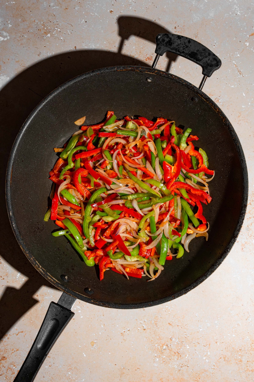 A wok with sliced green and red peppers and onions cooking in oil. The wok is on a tan counter.