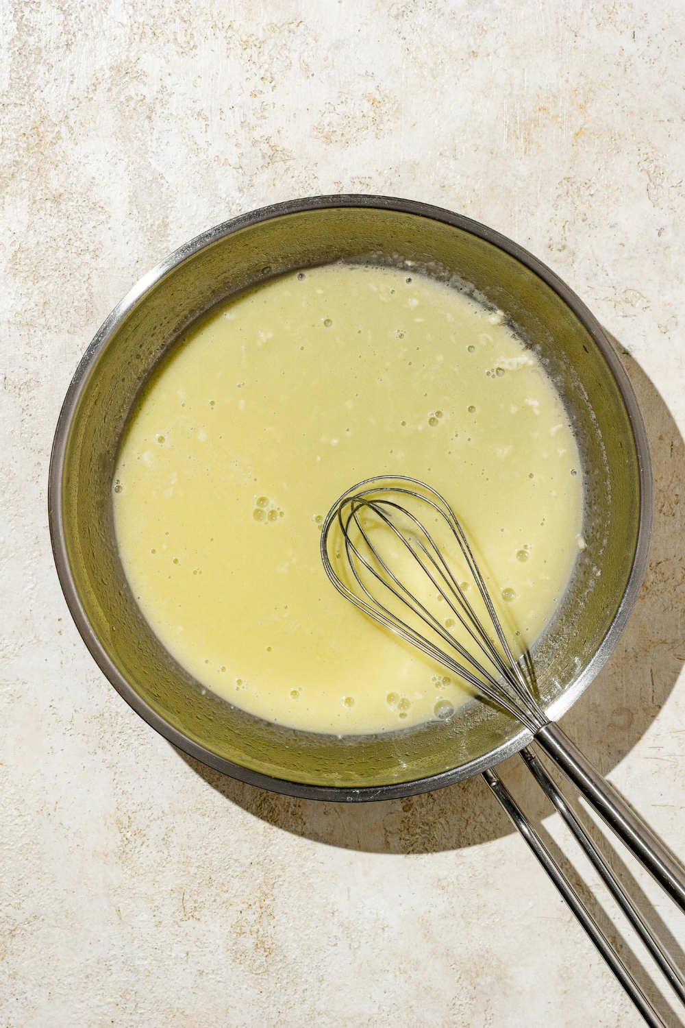 A skillet with a whisk stirring satuéed garlic and butter. The skillet is on a tan counter.