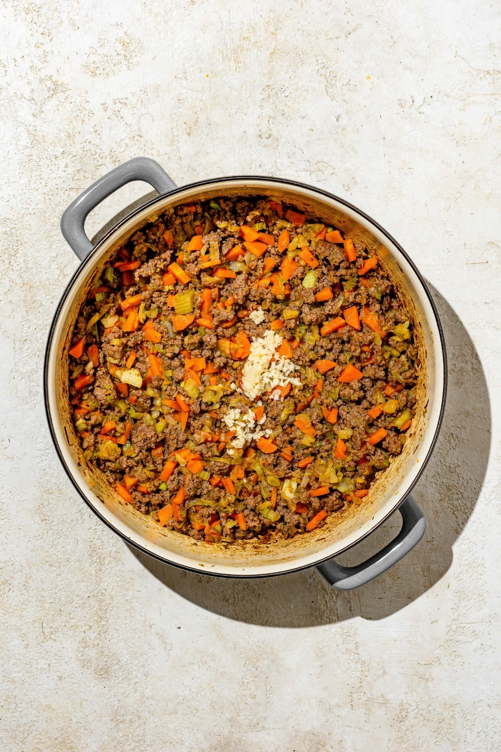 A dutch oven with ground pork, carrots, onions, celery, and seasonings cooking in oil. The pot is on a tan counter.