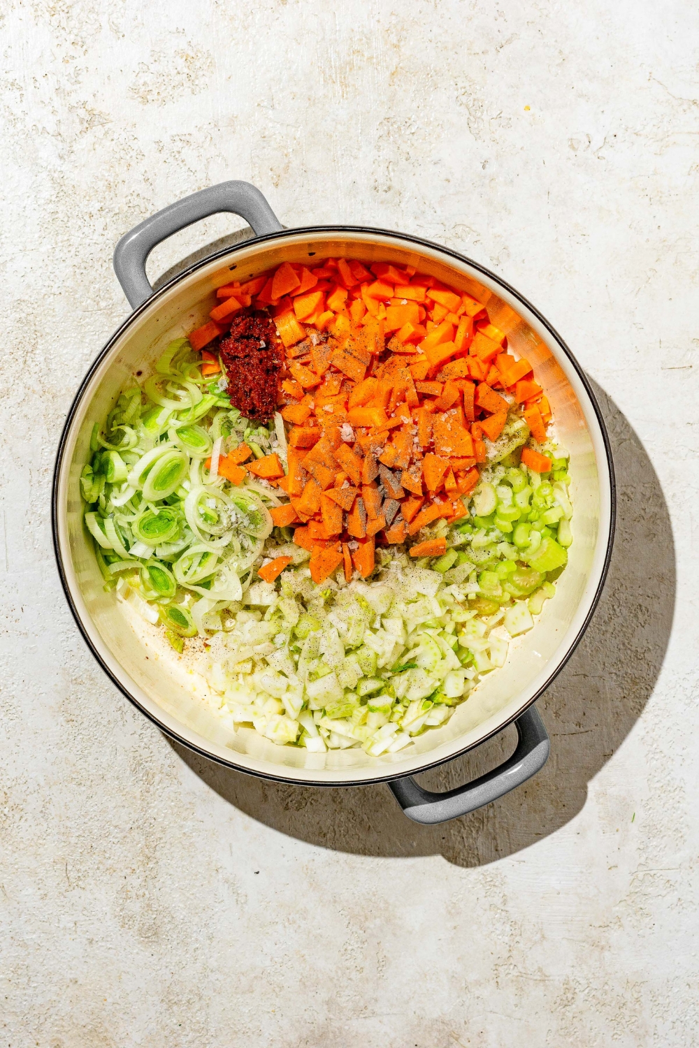 A dutch oven with diced carrots, onions, celery, leeks, and seasonings. The dish is on a tan counter.