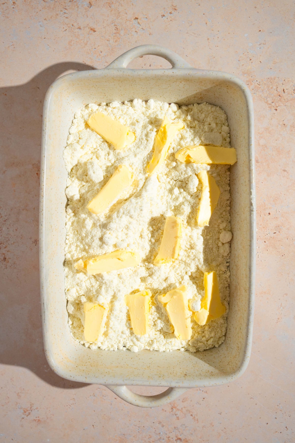 A baking dish with blueberry pie filling topped with vanilla cake mix and sliced butter. The baking dish is on a tan counter.