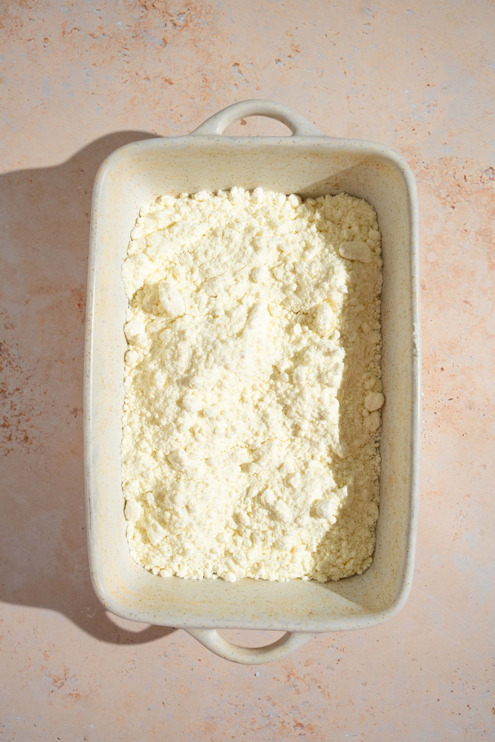 A baking dish with blueberry pie filling topped with vanilla cake mix. The baking dish is on a tan counter.