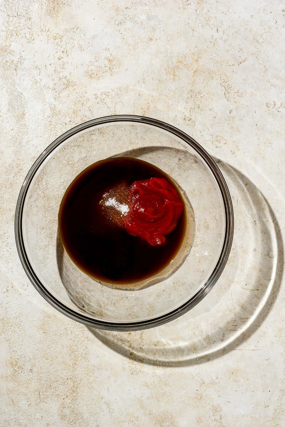 A glass bowl with ingredients to make chicken Katsu sauce sprinkled with salt. The bowl is on a tan counter.