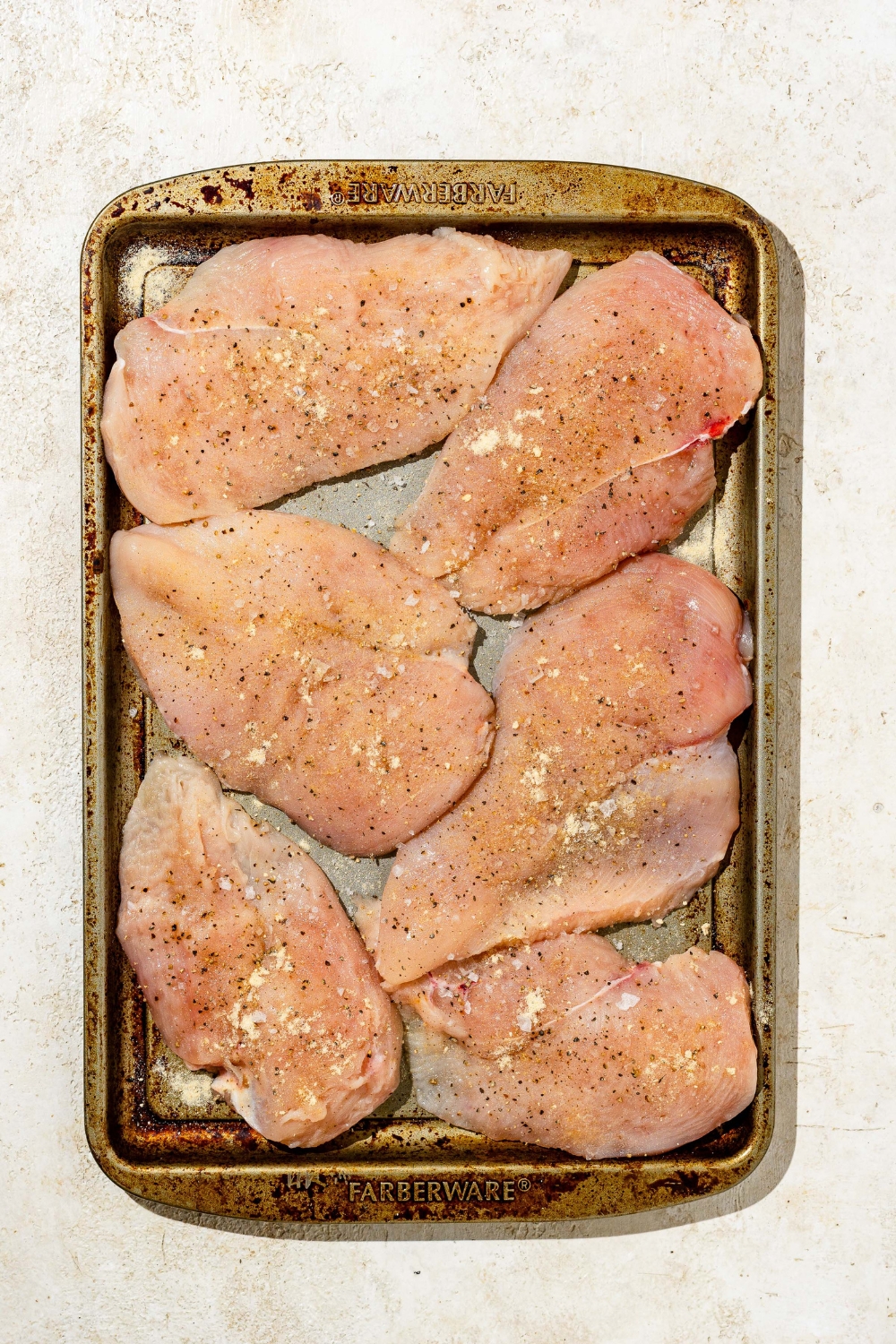 A baking sheet with several pieces of uncooked chicken breasts coated in seasonings. The sheet is on a tan counter.