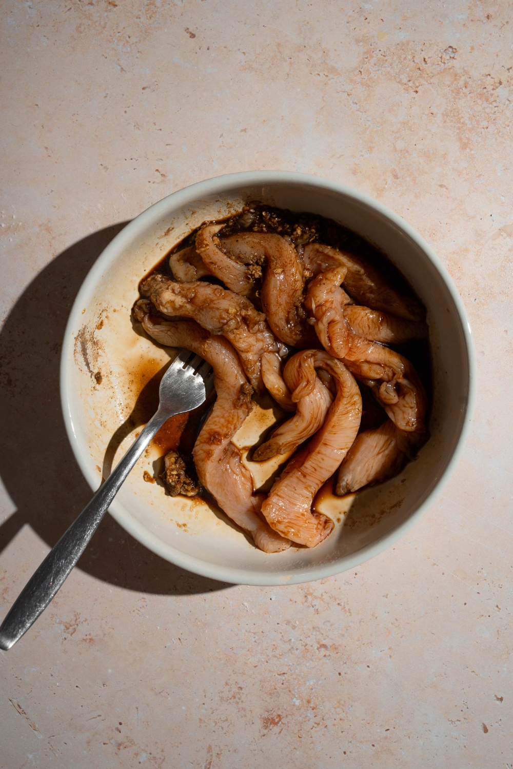 A white bowl with a fork with chicken marinating in a tempura marinade. The bowl is on a tan counter.