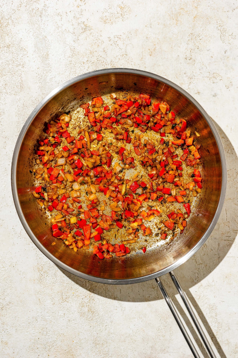 A skillet with diced pepper, onions, and seasonings cooking in oil. The skillet is on a tan counter.