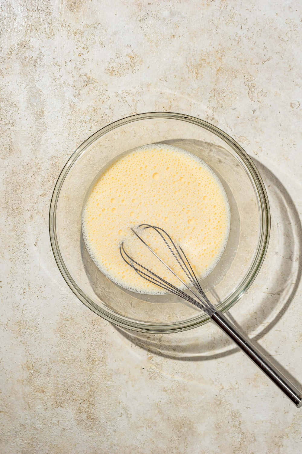A glass bowl with a whisk mixing an egg and milk mixture. The bowl is on a tan counter.
