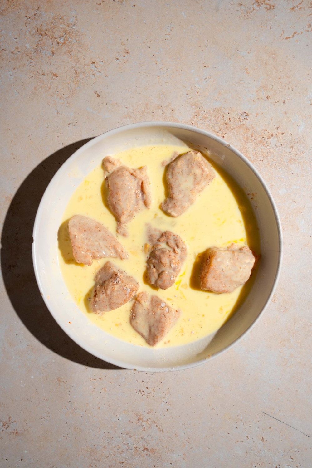 A bowl of egg batter with uncooked seasoned chicken nuggets. The bowl is on a tan counter.