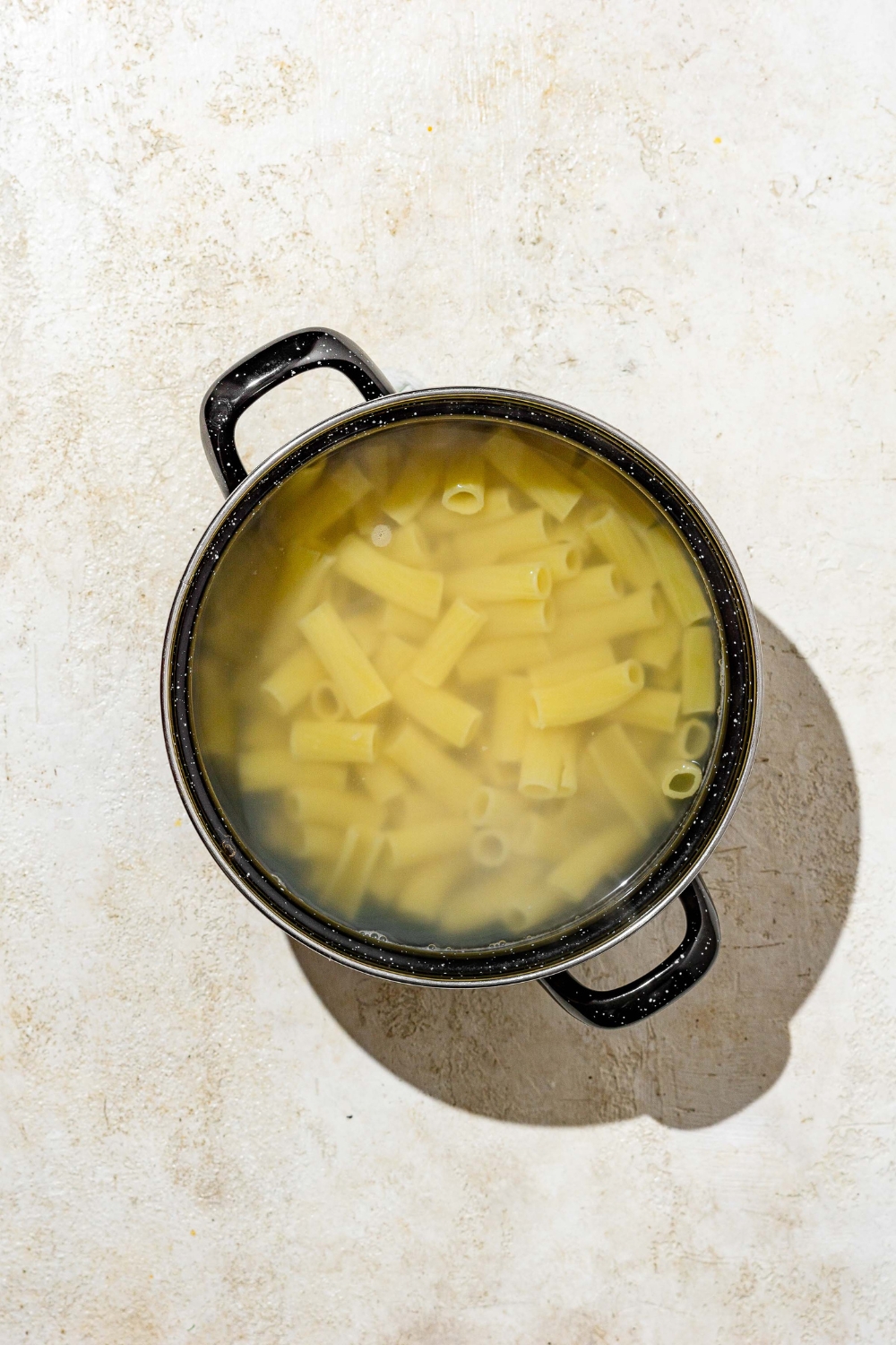 A stock pot with rigatoni pasta cooking in water. The pot is on a tan counter.