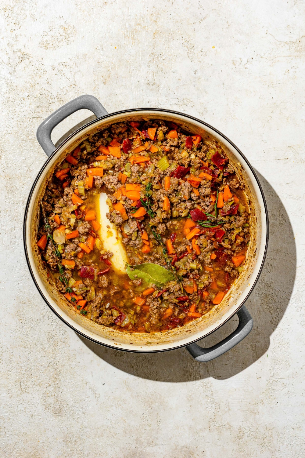 A dutch oven with a white bolognese base including ground pork, diced vegetables, and seasonings cooking in oil and white wine. The pot is on a tan counter.