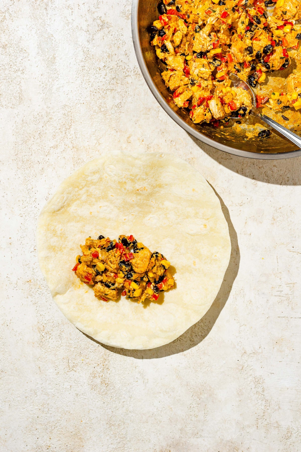 An open egg roll wrap with a southwestern chicken filling. The wrap is on a tan counter with a skillet of chicken mixture.