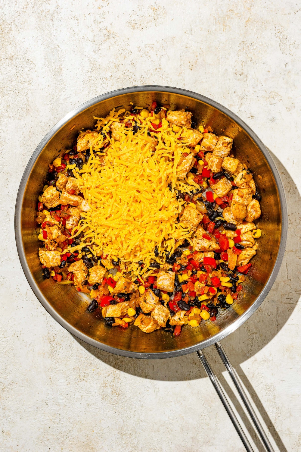 A skillet with cooked chicken, peppers, beans, seasonings, and corn topped with shredded cheese. The skillet is on a tan counter.