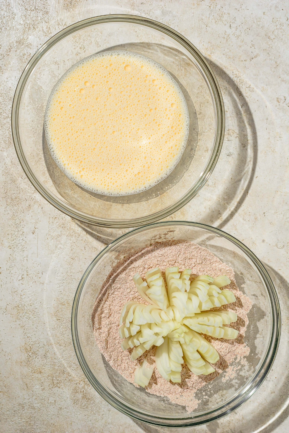 A glass bowl with an egg mixture next to a glass bowl with a bloomed onion in a flour and seasoning mixture.
