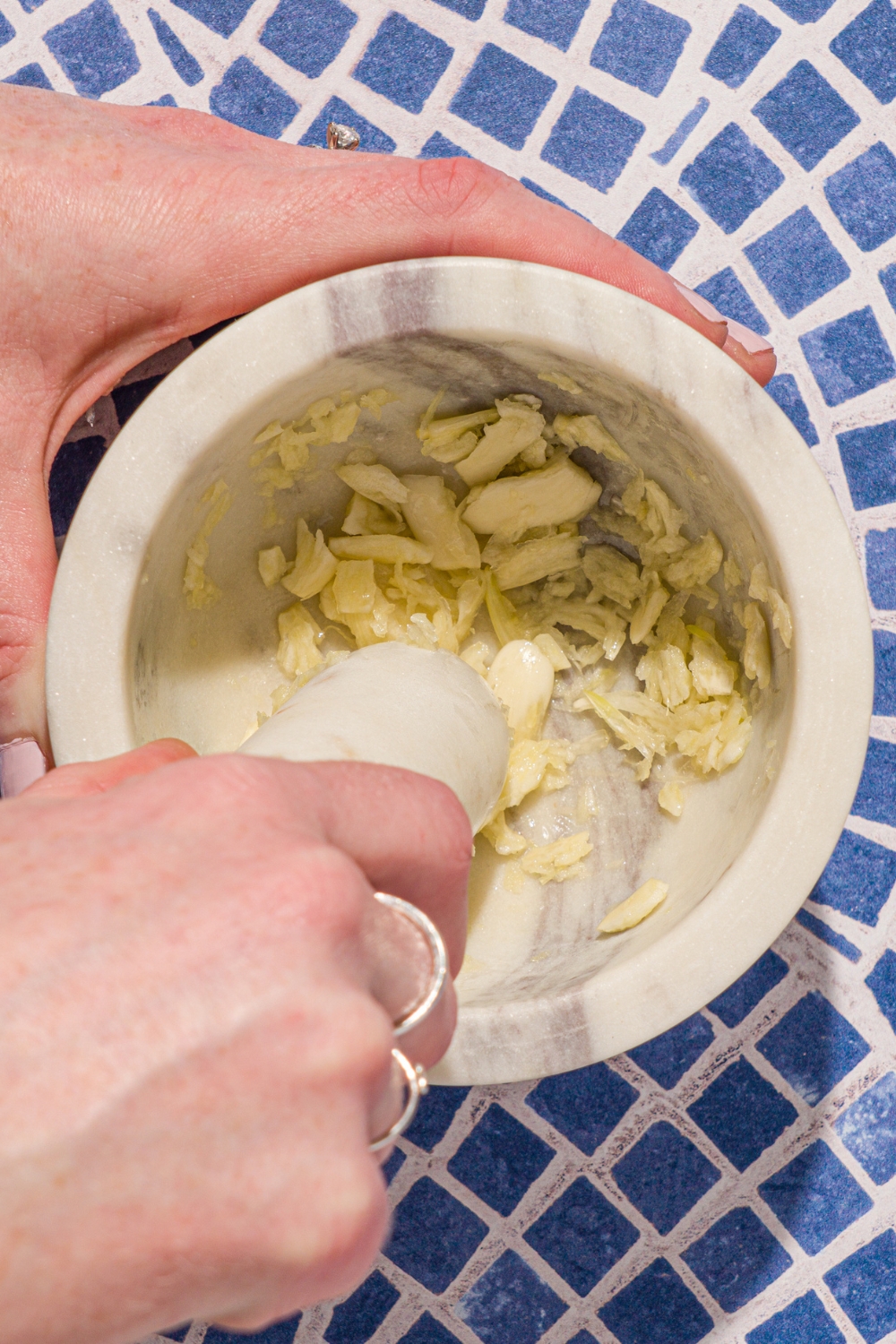 A hand using a mortar and pestle to mash garlic. The mortar and pestle is on a tiled counter.