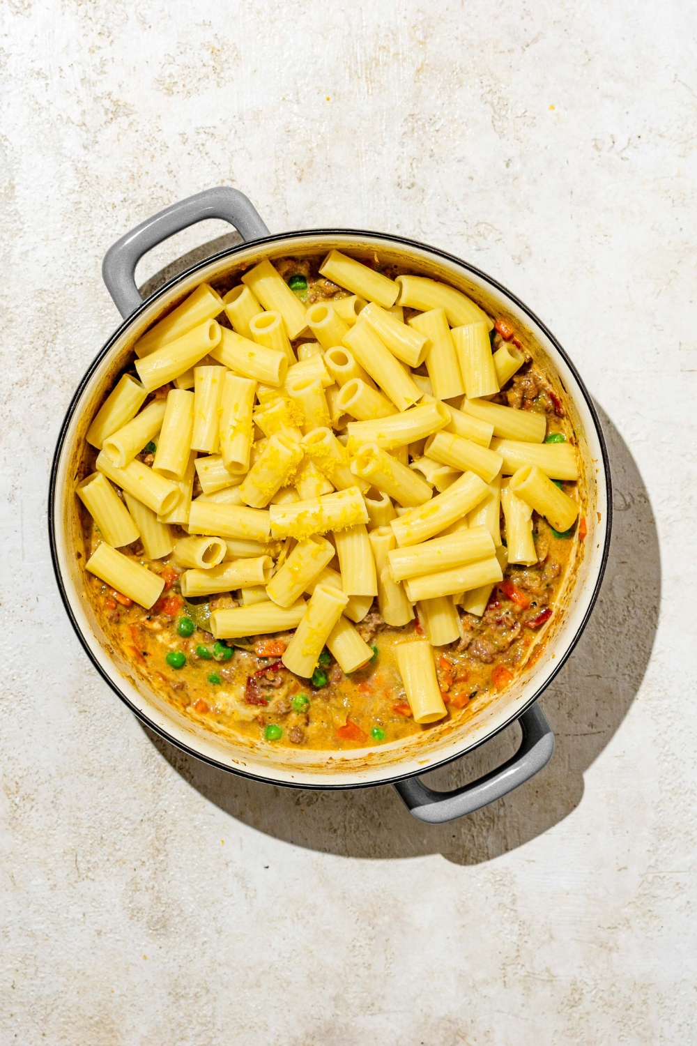 A dutch oven with white bolognese sauce topped with rigatoni pasta. The pot is on a tan counter.
