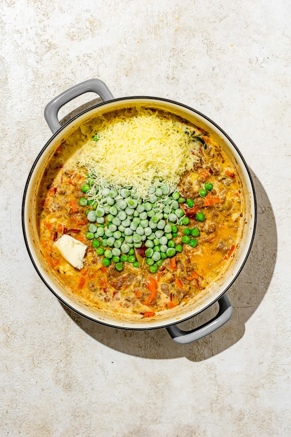 A dutch oven with white bolognese sauce topped with grated parmesan cheese, frozen peas, and butter. The pot is on a tan counter.
