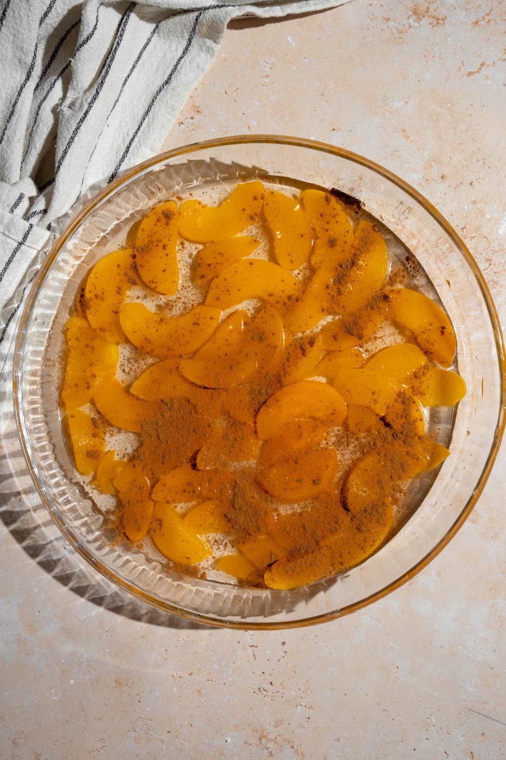 A glass baking dish with canned peaches topped with cinnamon. The dish is on a tan counter with a white striped napkin.
