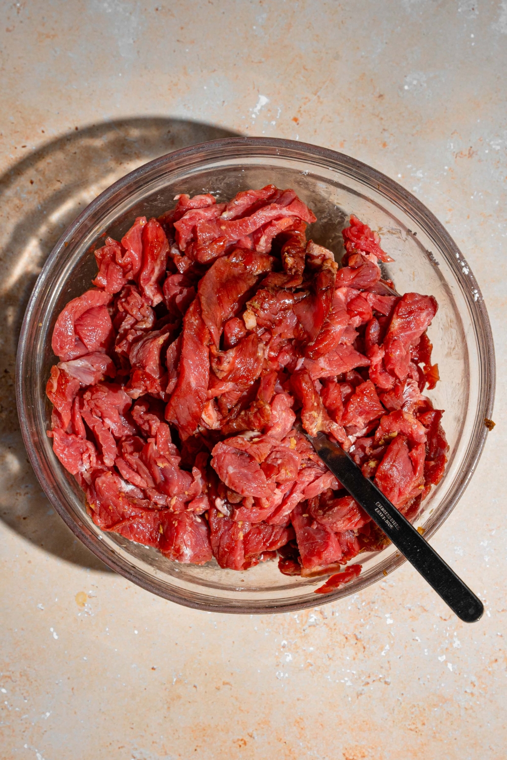 A glass bowl with sliced uncooked steak tossed in marinade. There is a spoon in the bowl.