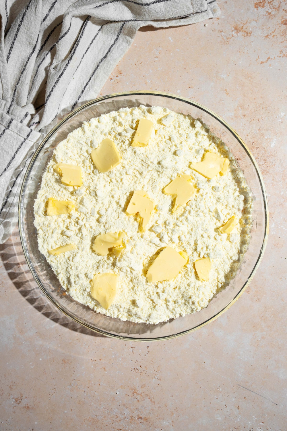 A glass baking dish with layered ingredients to make lemon cream cheese dump cake including lemon filling, cream cheese, cake mix, and butter. The dish is on a tan counter.