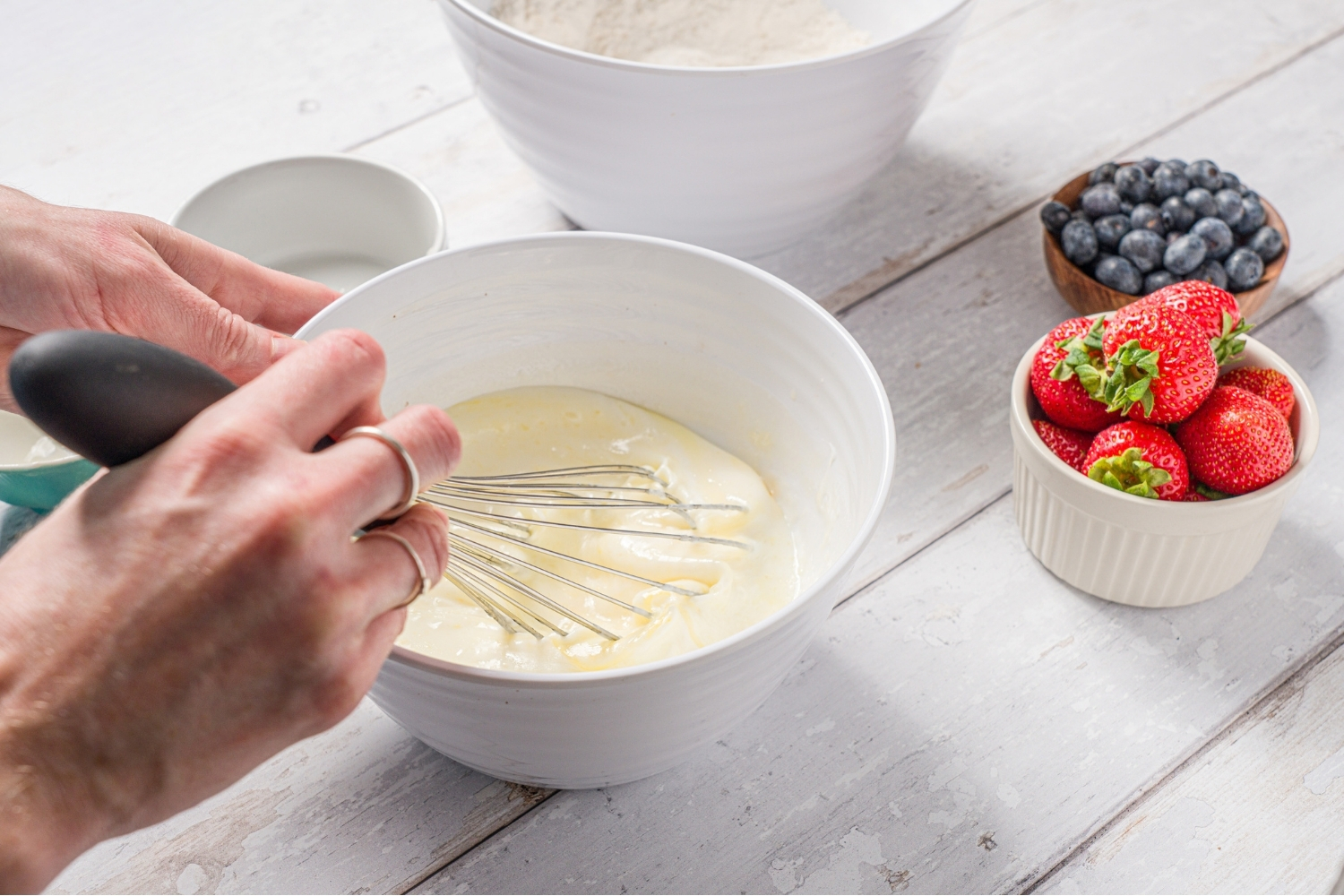 A white bowl of a hand whisking wet ingredients to make reindeer pancakes. The bowl is on a wood counter with a bowl of dry ingredients and two bowls of berries.