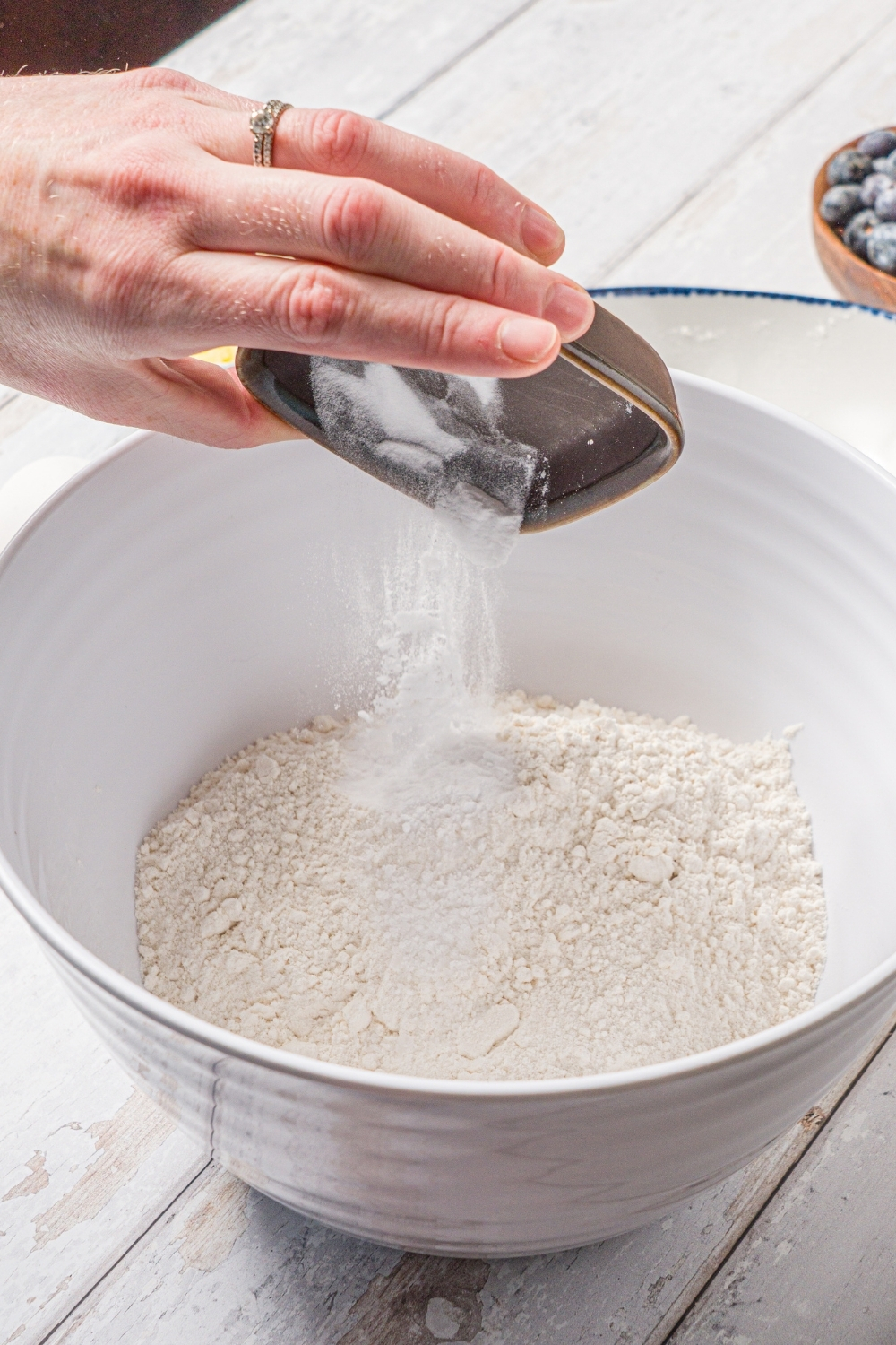 A white bowl with dry ingredients to make reindeer pancakes including flour. A hand is dumping a small bowl of baking soda into the mixture. The bowl is on a wood counter.