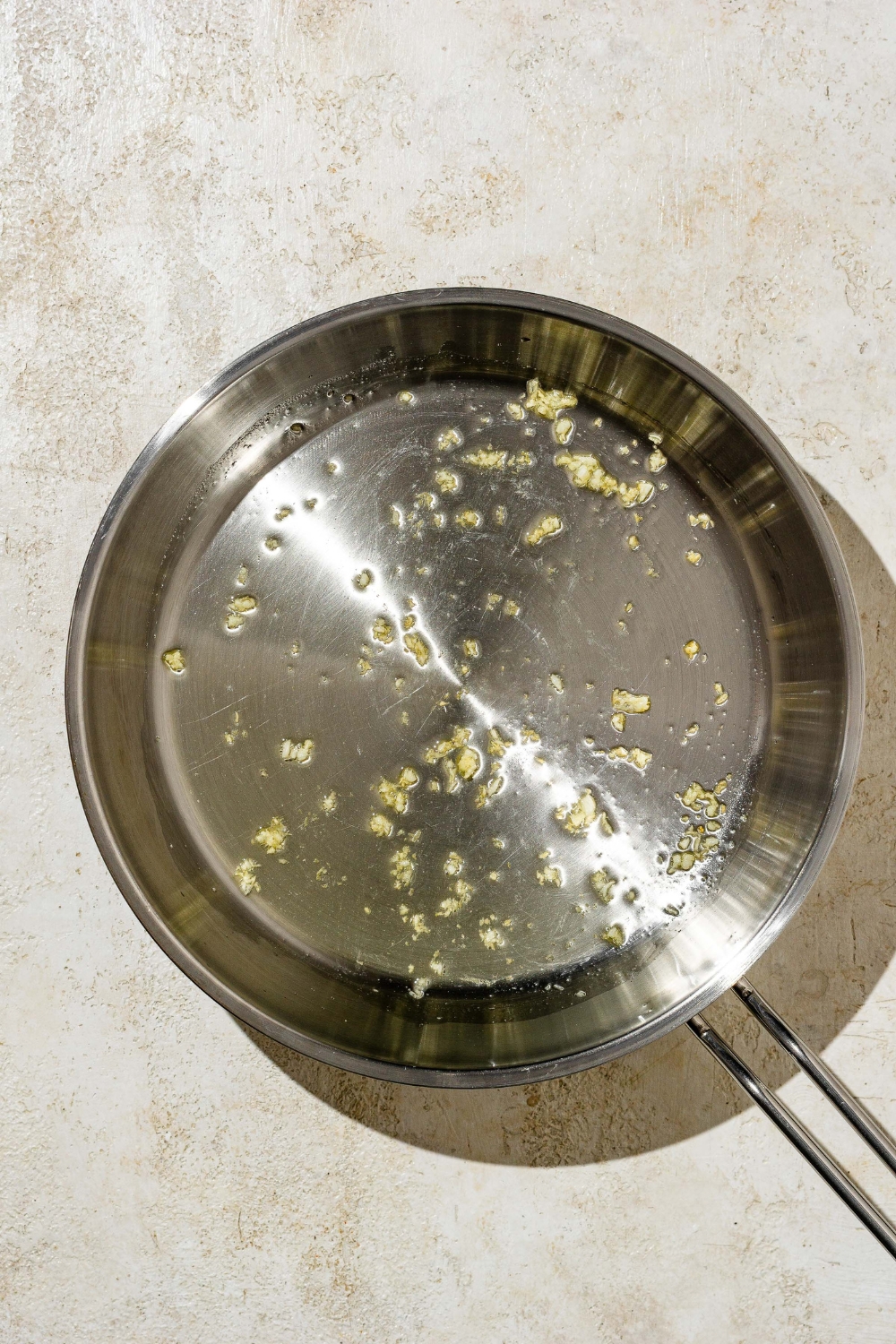 A skillet with garlic cooking in oil. The skillet is on a tan counter.