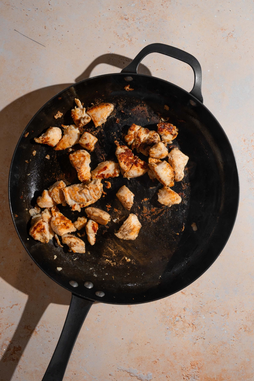 A skillet with pieces of chicken sautéed in oil. The skillet is on a tan counter.