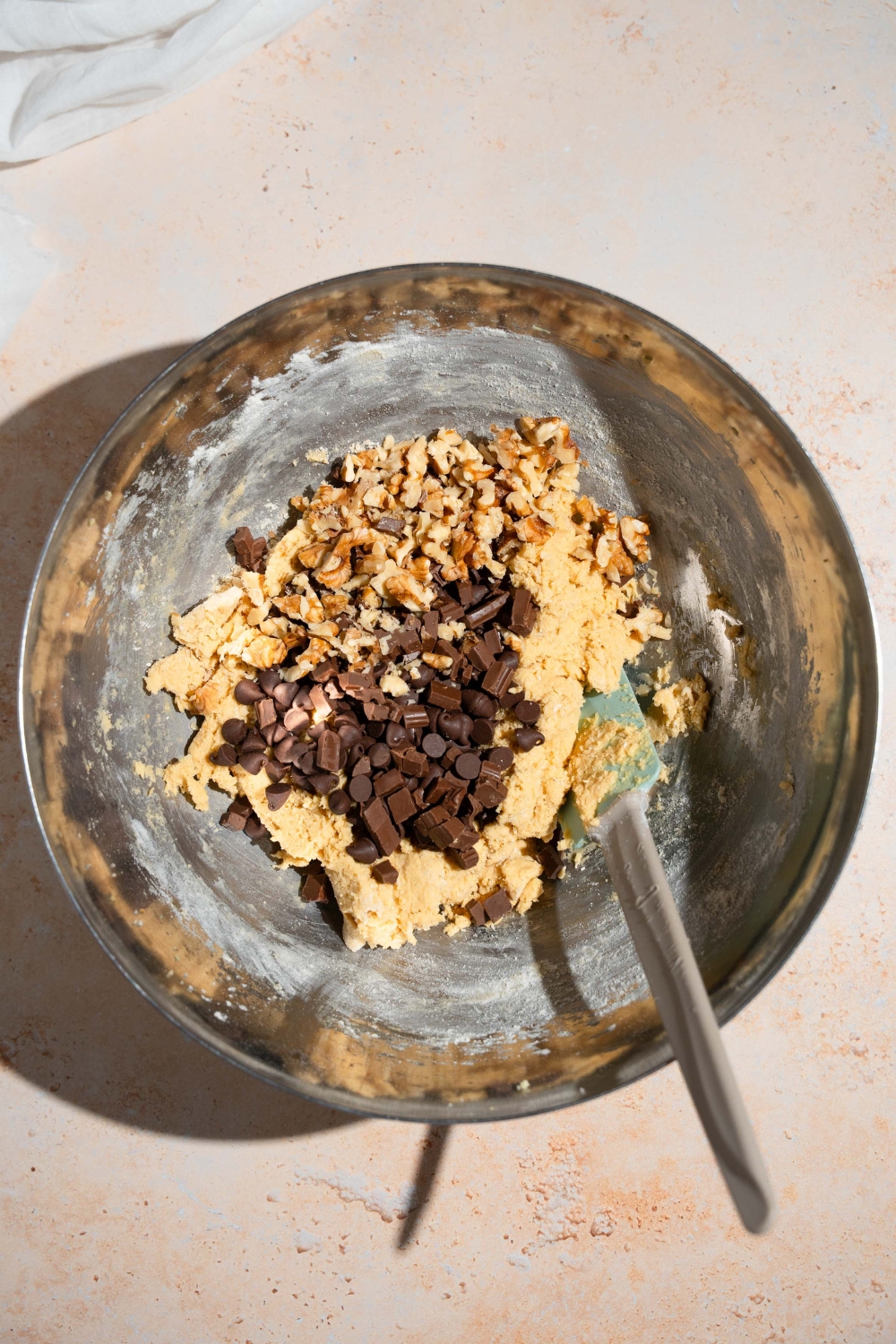 A mixing bowl with a spatula mixing chocolate chips and walnuts into a cookie dough mixture. The bowl is on a tan counter.