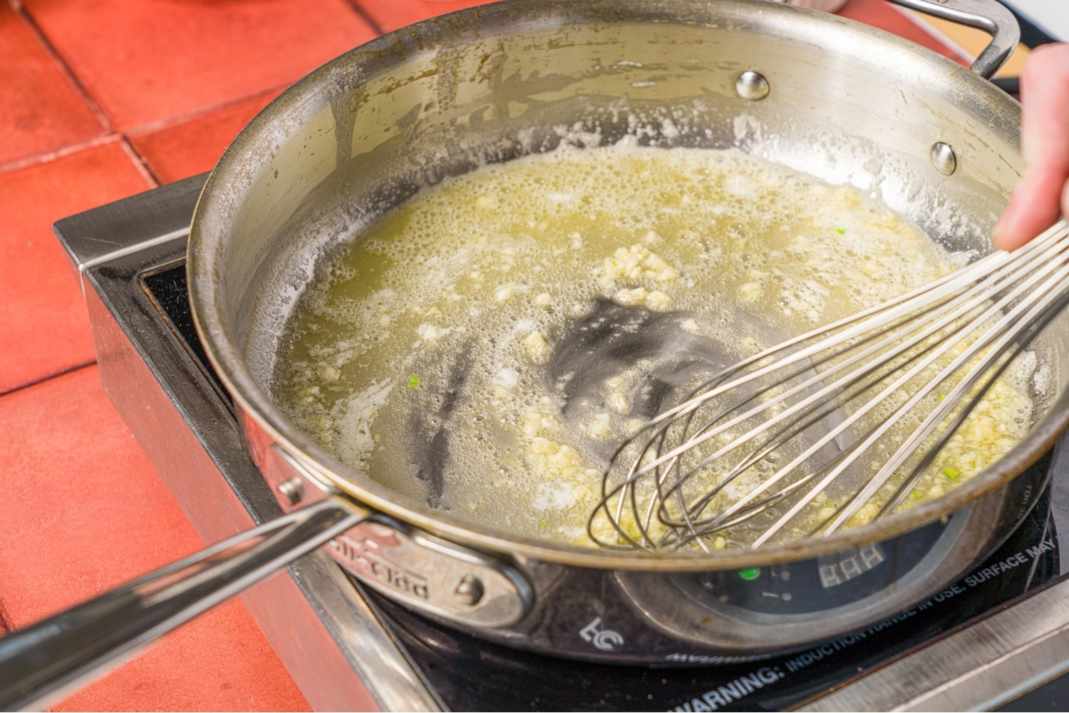 A skillet with melted butter and garlic cooking on a burner. There is a hand stirring the butter with a whisk. The burner is on a brick counter.